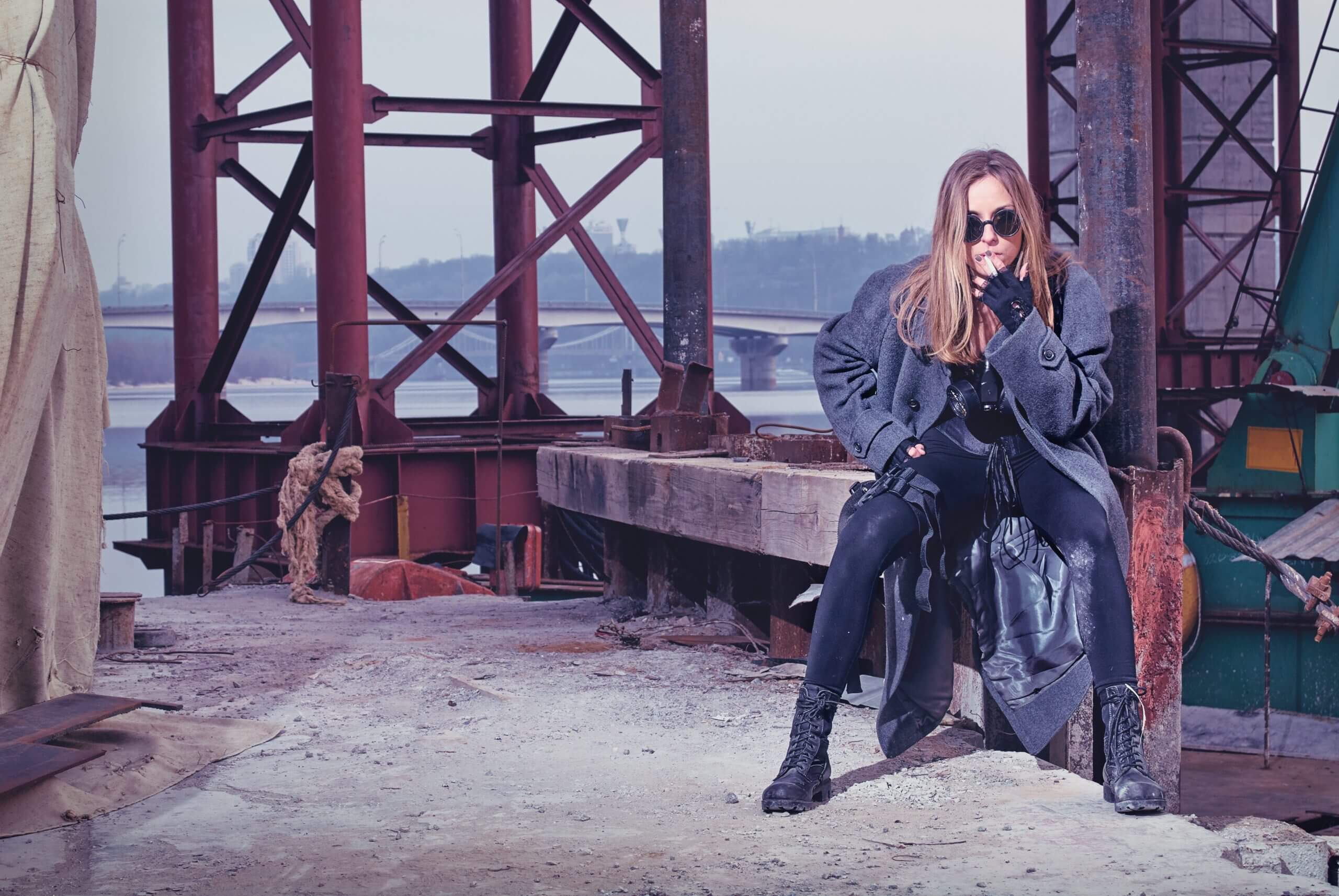 Woman in trench coat, dark clothing and sunglasses waiting for someone under a bridge