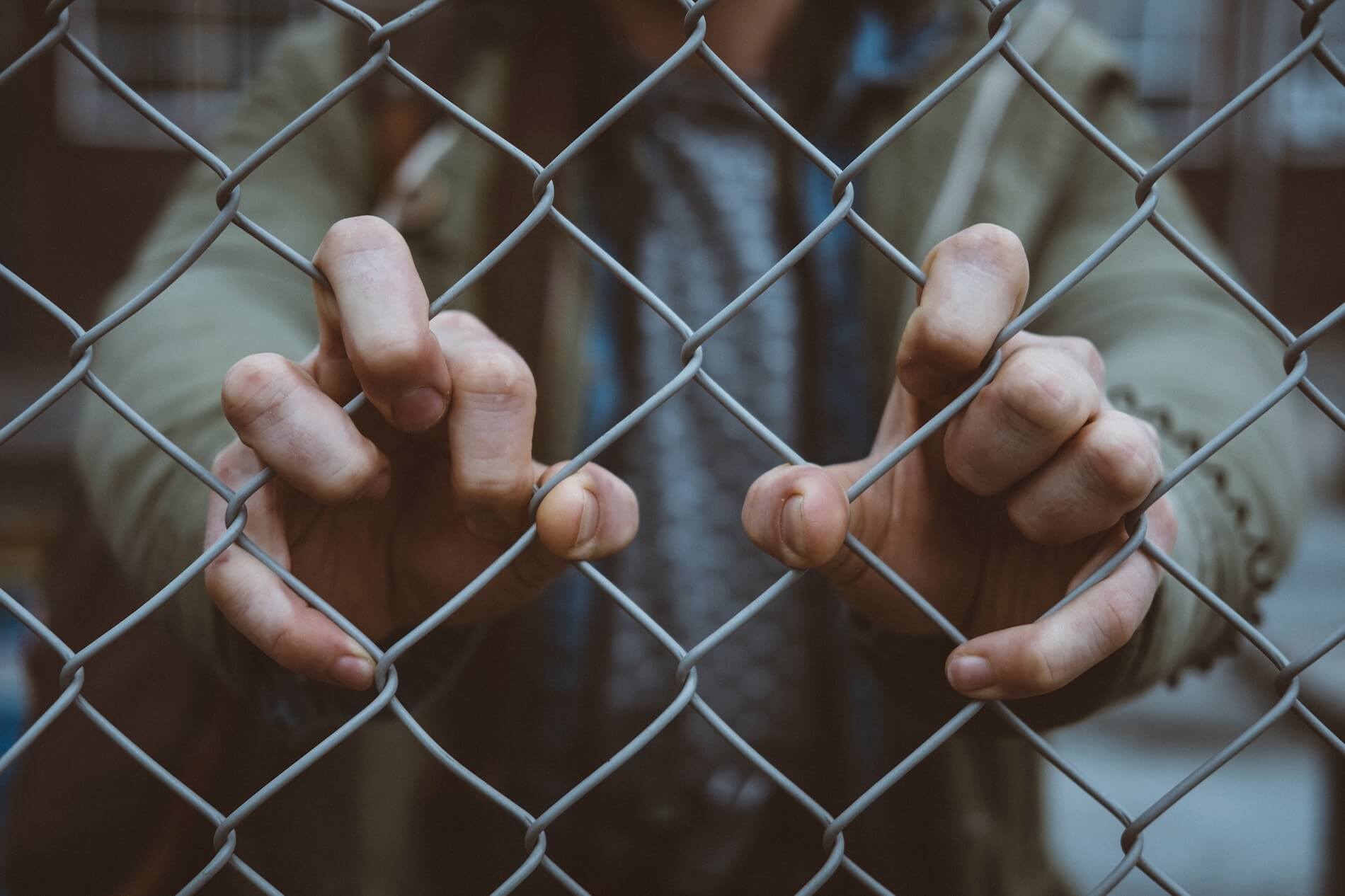 Blurry image of a torso in a beige jacket with his fingers gripping chain link fencing