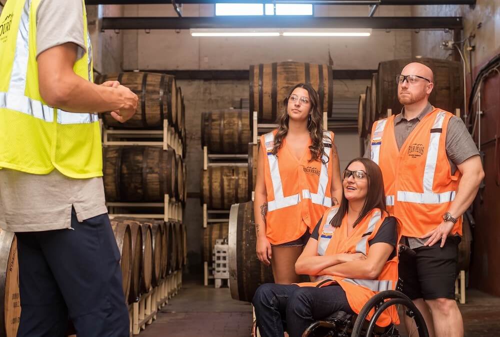 Four individuals in a warehouse setting wearing brightly colored safety vests, one of them a woman in a wheelchair