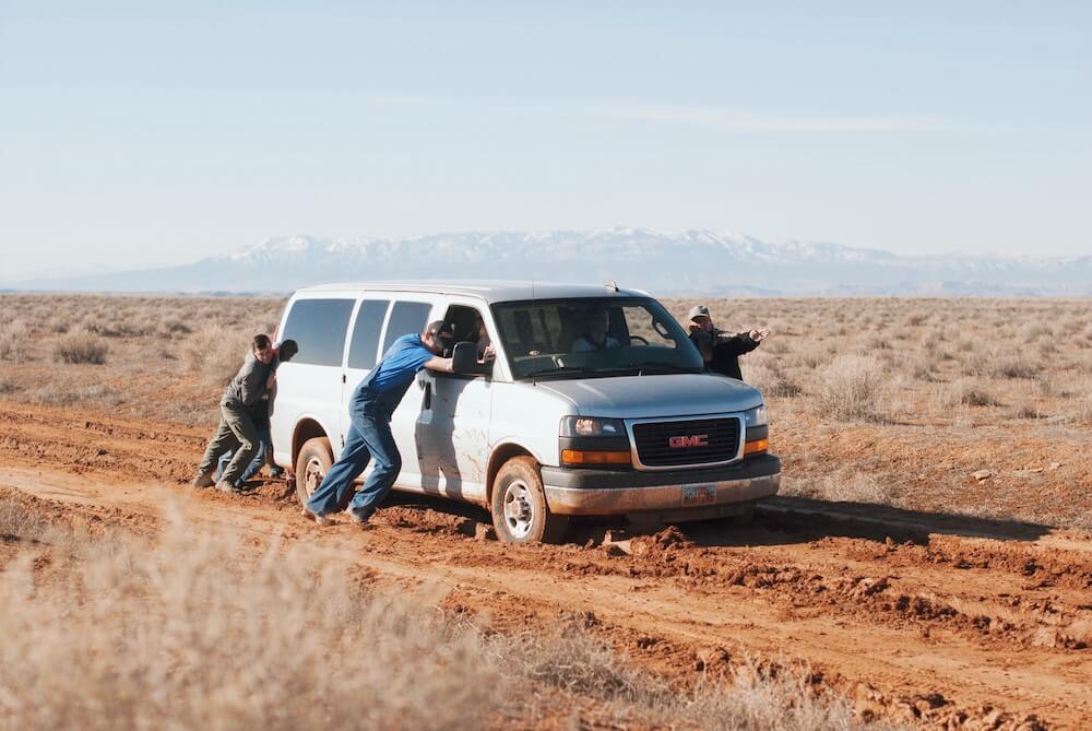 Old white econoliner-type van stuck in the mud in a remote area being pushed by three people