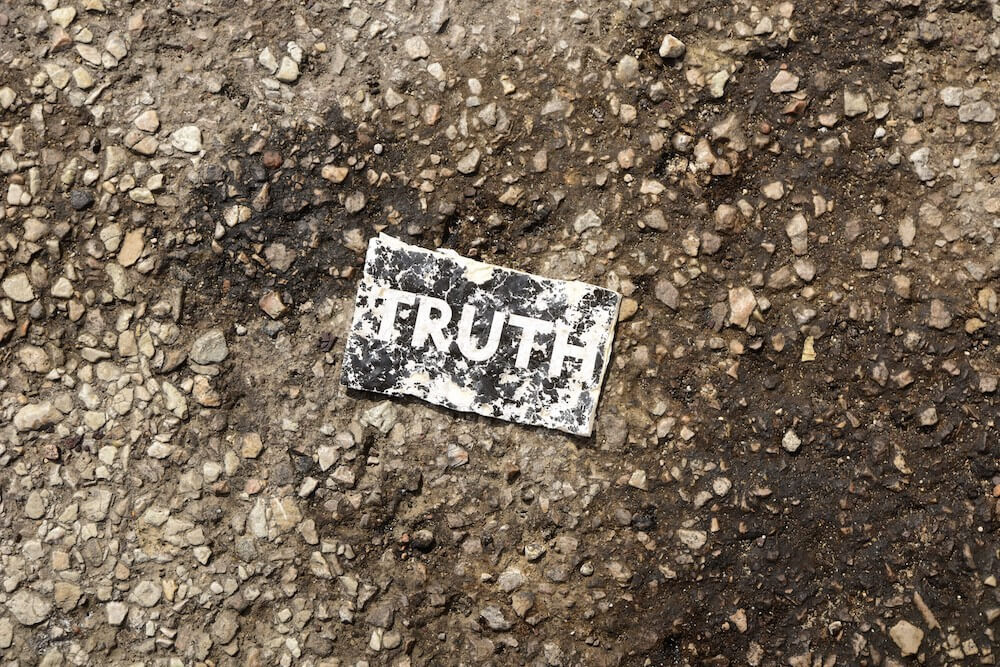 Worn black piece of paper with white letters spelling TRUTH in a pothole on worn asphalt