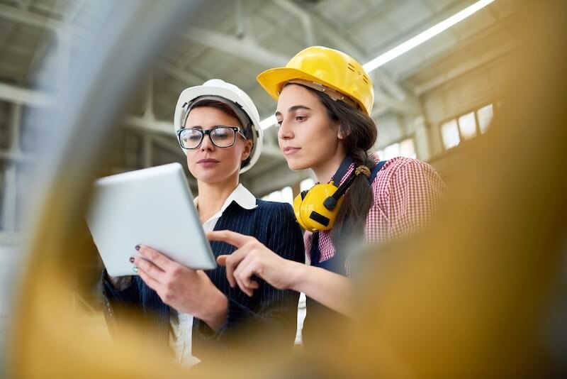 Two women in hard hats looking at a tablet in a warehouse setting